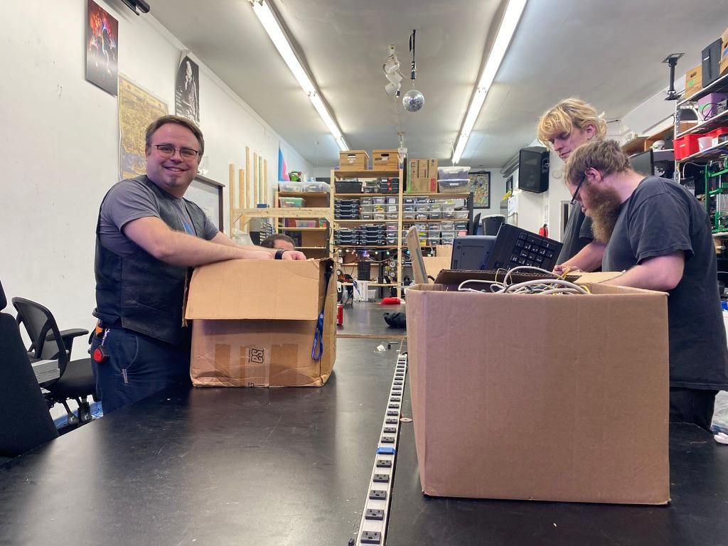 Three people standing in front of boxes of used electronics on a table in a brightly lit room full of electronics.